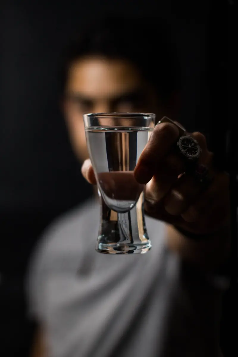 Greek sosteniendo un caballito de tequila frente a la cámara, en primer plano desenfocado