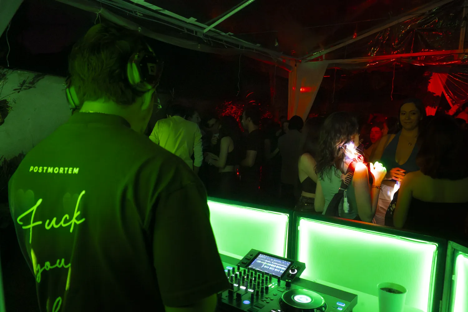 Greek de espaldas en la cabina, con barra iluminada en verde y público al fondo
