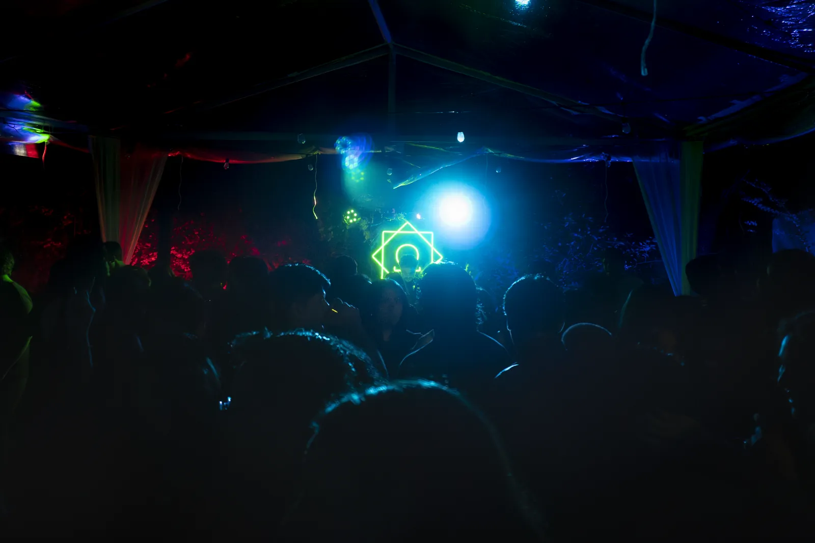 Público en una fiesta frente a la cabina, con el letrero de neón Ω al fondo bajo luces azules